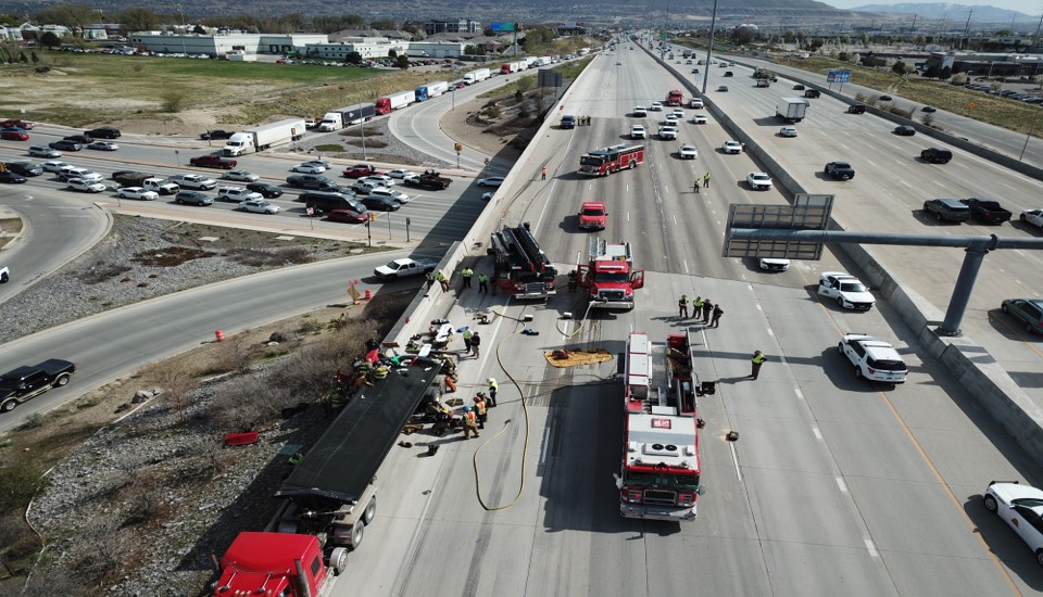 A closed section of interstate highway passes over a busy roadway. Emergency response vehicles surround a tractor trailer.